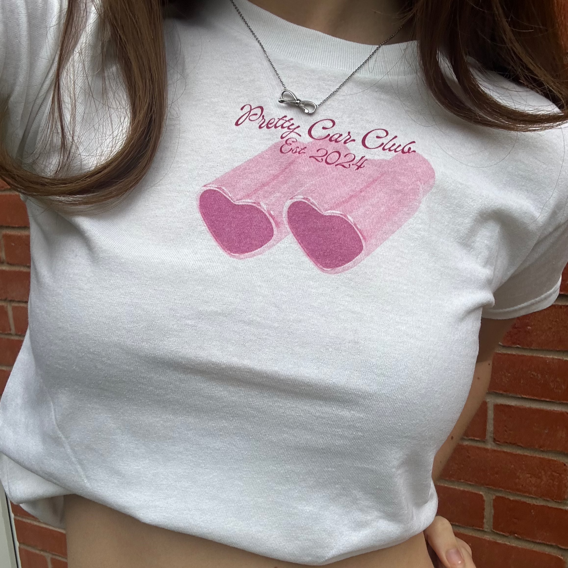 White baby tee with a small pink design in the centre. The design consists of two heart shaped exhausts beneath the text Pretty Car Club Est 2024. Perfect for car enthusiasts looking for feminine car merchandise. 